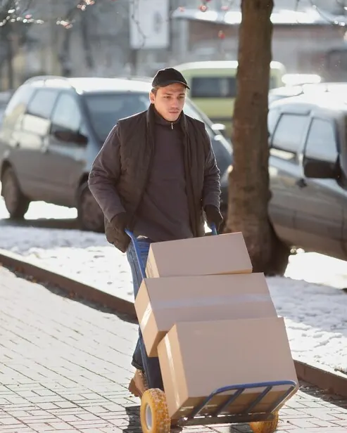 Curier care împinge pe trotuar un cărucior cu trei cutii, pe o stradă cu mașini parcate